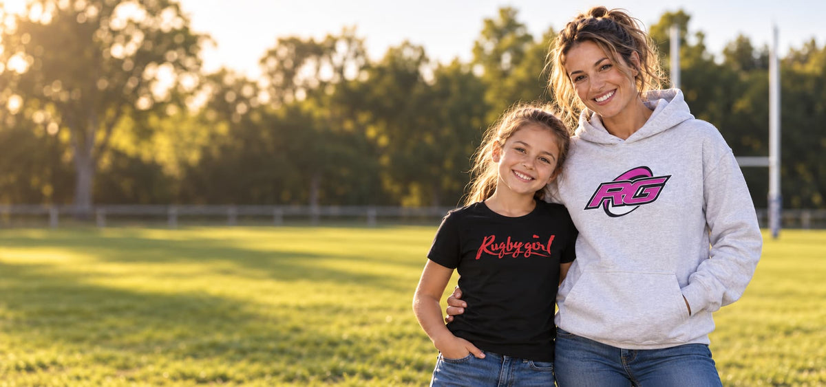 A woman and a young girl standing together on a grassy field at sunset. The woman wears a light gray hoodie with a pink and black 'RG' logo, and the girl wears a black t-shirt with a red 'Runbranded' logo.